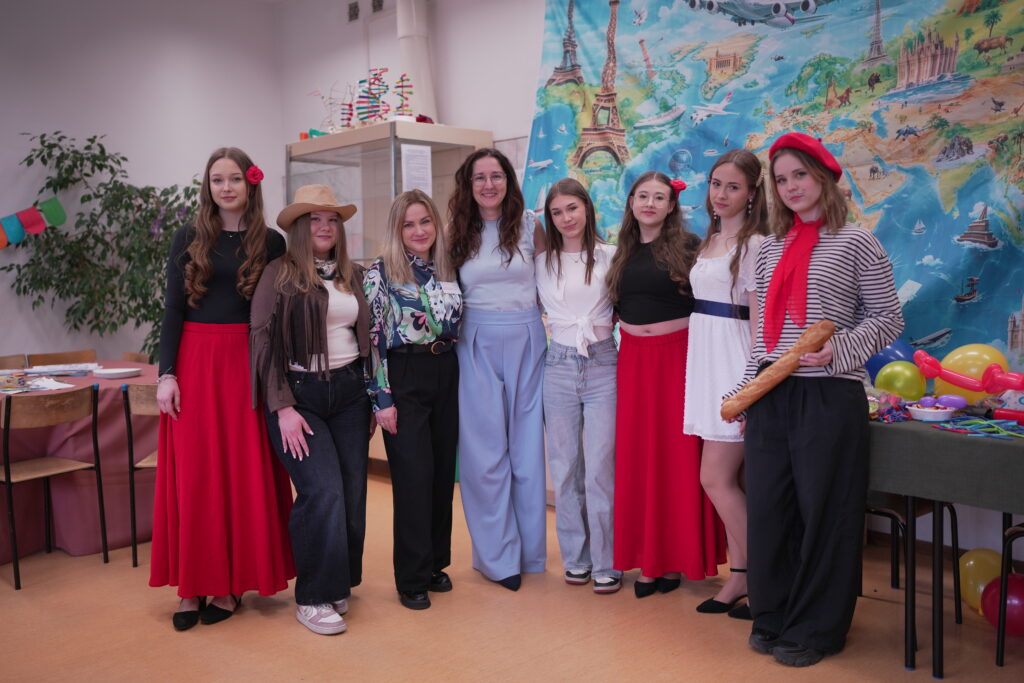 Grupa młodych kobiet ubranych w różnorodne stroje, w tym w stylu francuskim, pozująca na tle kolorowej mapy świata w klasie.