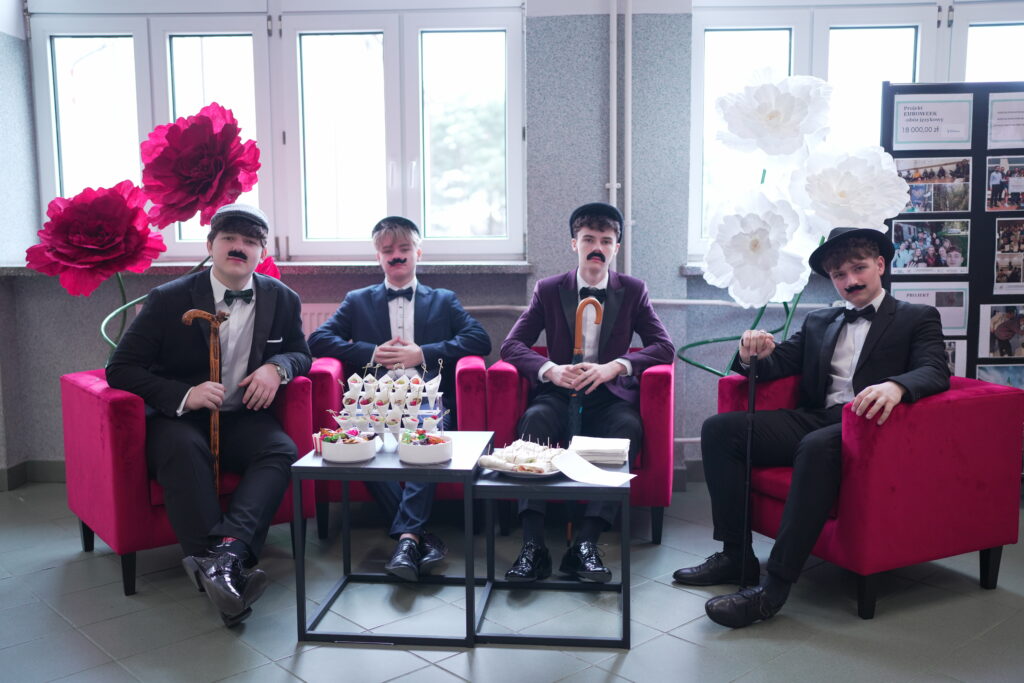 Czterech młodych mężczyzn w eleganckich garniturach, w melonikach i z fałszywymi wąsami, siedzi na czerwonych fotelach obok stolików z przekąskami.