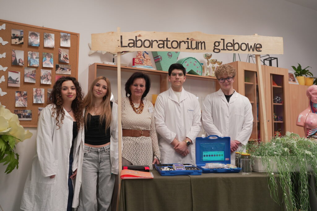 Grupa osób stojąca przy stanowisku z napisem "Laboratorium glebowe", na stole znajdują się profesjonalne zestawy do badań gleby oraz zielone rośliny.