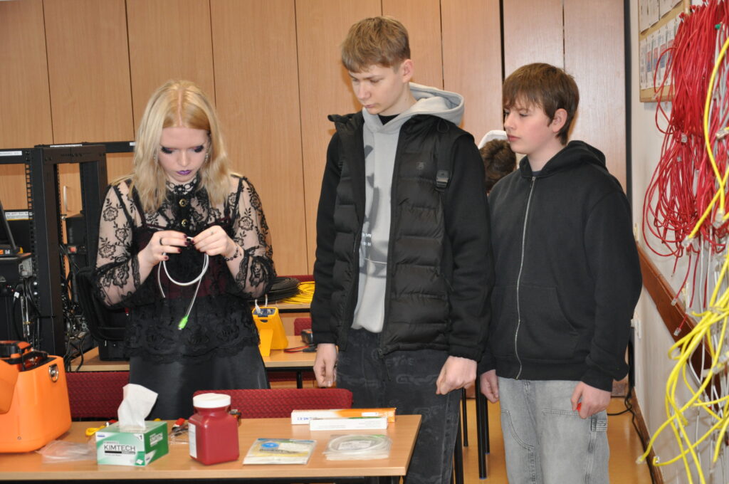 Trójka młodzieży stoi przy stole z narzędziami i kablami, dziewczyna skupiona na pracy z kablem, dwóch chłopców obserwuje ją z boku.