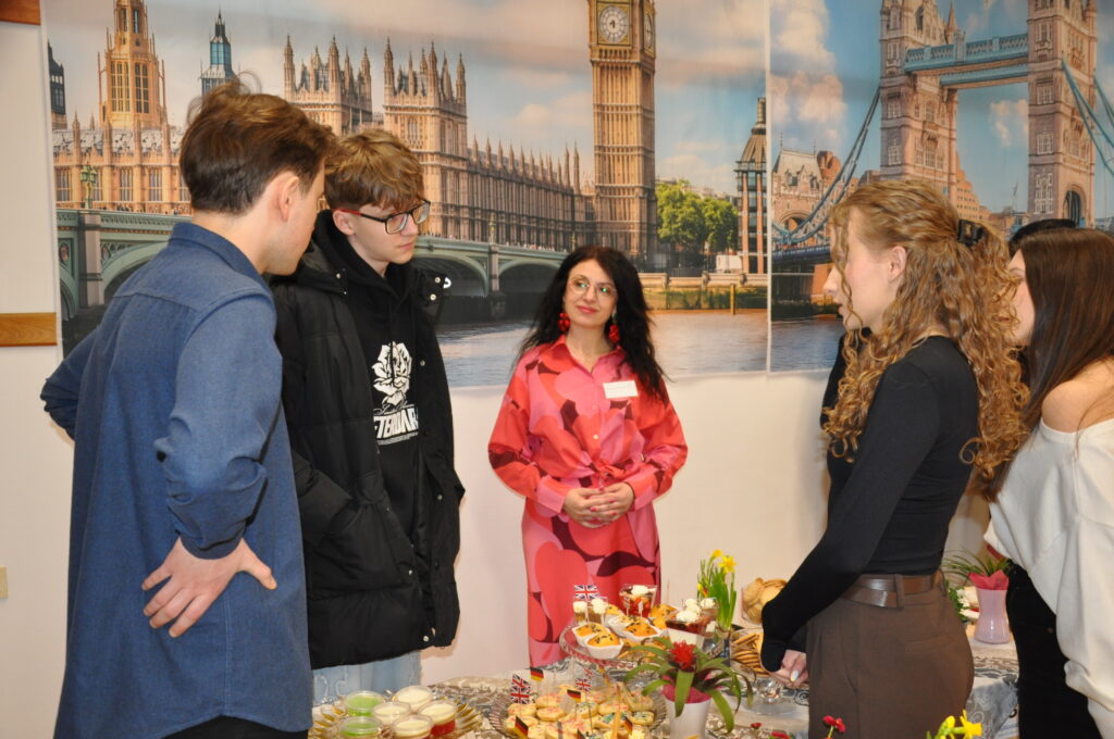 Grupa młodych ludzi rozmawia przy stole z różnorodnymi przekąskami, na tle dekoracji z motywem Londynu, w tym Big Bena i Tower Bridge.