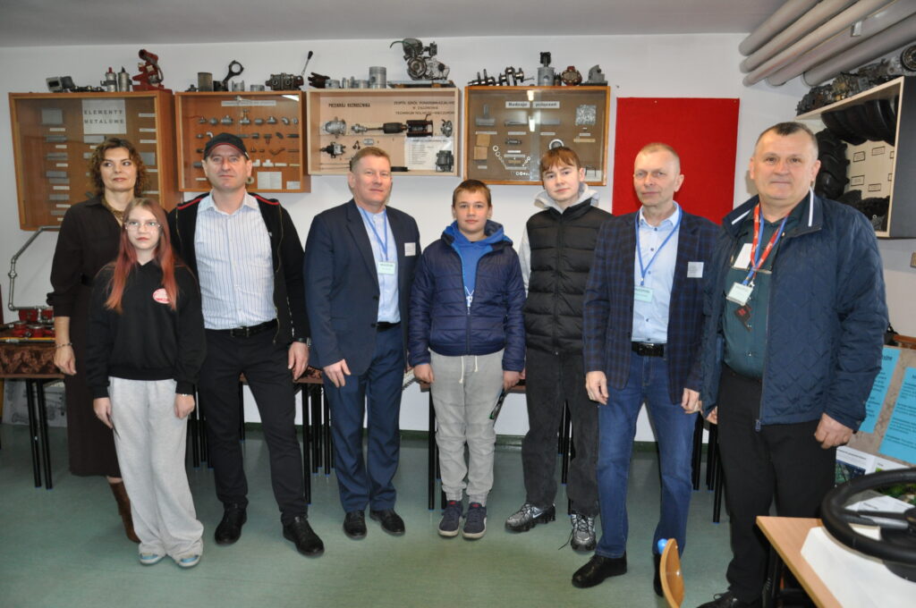 Grupa ludzi w różnym wieku stoi w sali edukacyjnej z wystawionymi metalowymi częściami i tablicami informacyjnymi dotyczącymi techniki rolnej.