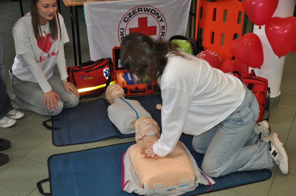 Kobieta ćwiczy resuscytację na fantomie, a obok siedzi druga kobieta obok sprzętu medycznego i balonów w kształcie serc.