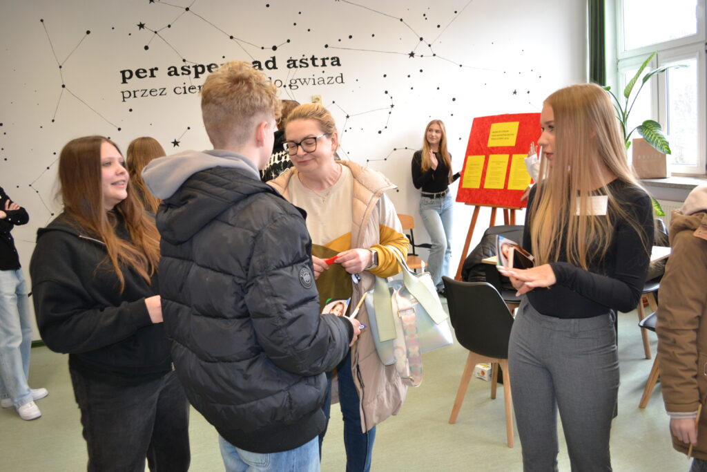 Grupa młodych osób rozmawia w jasnym pokoju z napisem "per aspera ad astra przez ciernie do gwiazd" na ścianie, przy planszy z informacjami.