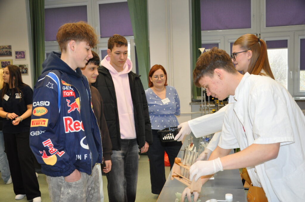 Grupa młodzieży obserwuje doświadczenie chemiczne przeprowadzane przez uczniów w białych fartuchach i rękawiczkach.