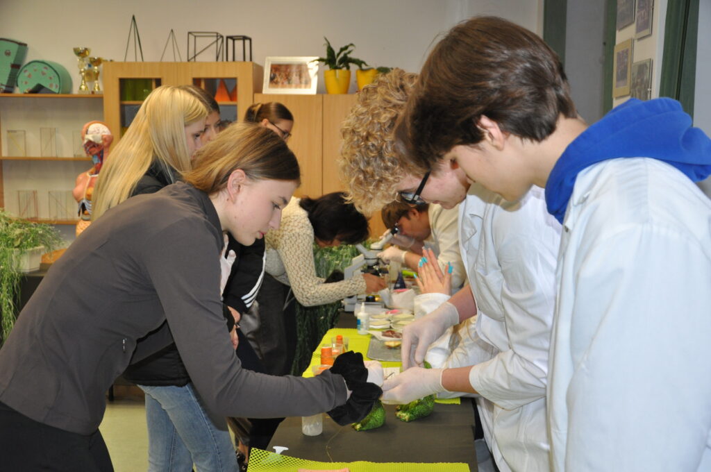 Grupa młodych osób w laboratorium biologicznym z białymi fartuchami i rękawiczkami, przeprowadzająca doświadczenia na zielonych próbkach.