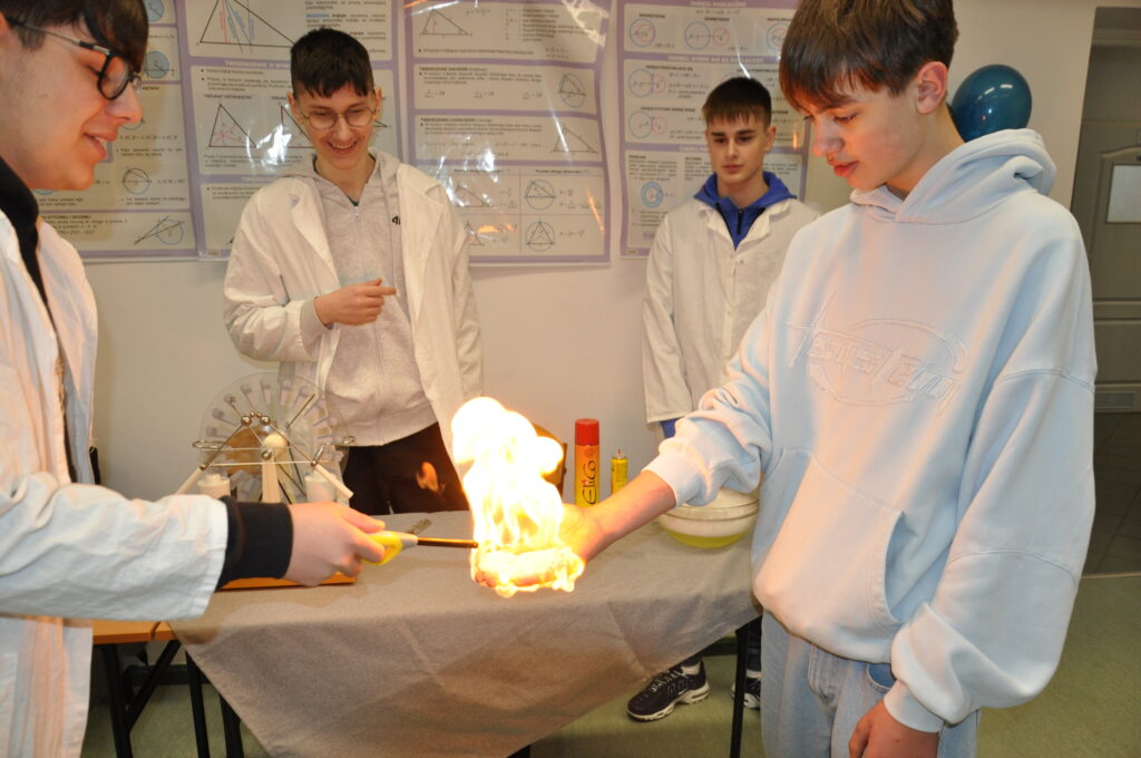 Czterech nastolatków obserwuje eksperyment z ogniem, podczas którego jeden z nich trzyma płonącą rękę przykrytą specjalną substancją, a drugi podpala ją zapalniczką.