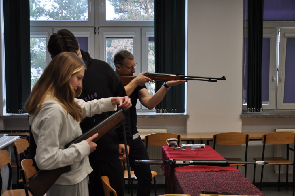 Trzy osoby ćwiczą strzelanie z wiatrówek w pomieszczeniu przypominającym salę lekcyjną, jedna osoba mierzy do celu, a dwie pozostałe przygotowują broń.