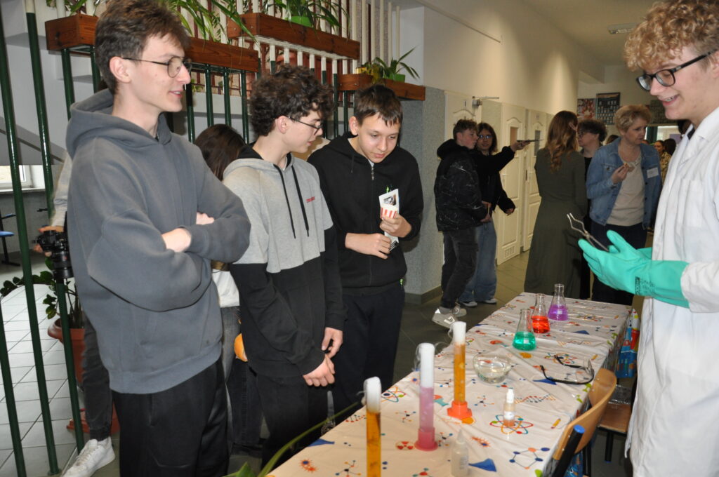 Grupa nastolatków obserwuje eksperyment chemiczny przy stole zkolorowanymi płynami i sprzętem laboratoryjnym, prowadzony przez chłopaka w białym fartuchu i zielonych rękawicach.