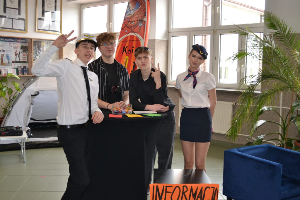 Grupa młodych ludzi stoi przy stole z napisem „INFORMACJA”, dwie osoby są ubrane w stroje lotnicze z białymi koszulami i krawatami.