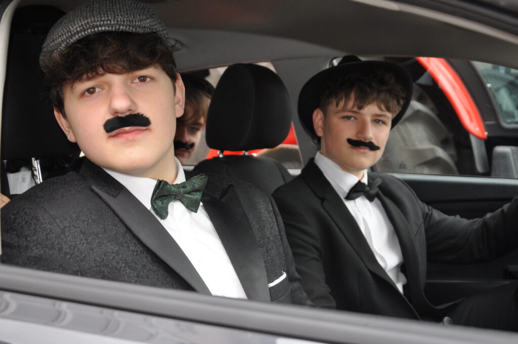 Trzech młodych mężczyzn w garniturach i sztucznych wąsach siedzi w samochodzie, dwaj z nich mają kapelusze.