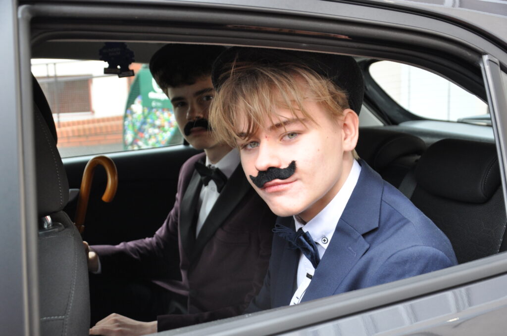 Dwaj młodzi mężczyźni w garniturach z przylepionymi sztucznymi wąsami siedzą w tylnej części samochodu.