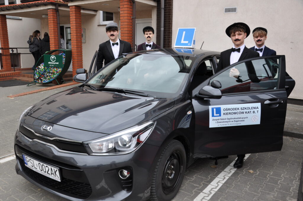 Czterech młodych mężczyzn w strojach retro z sztucznymi wąsami stoi przy samochodzie szkoły nauki jazdy z otwartymi drzwiami.