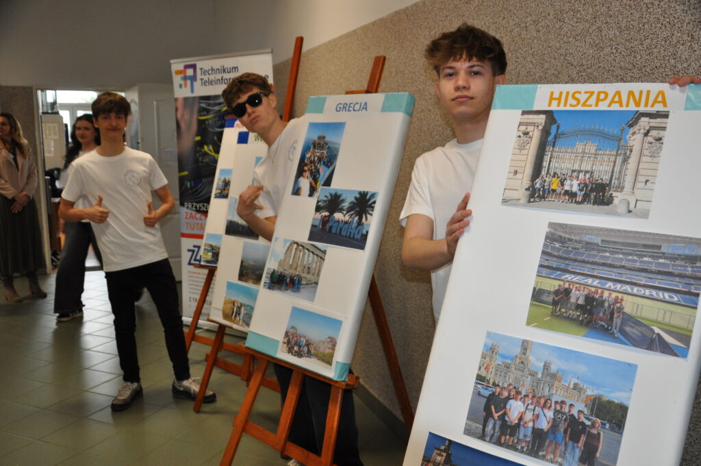 Trzech chłopców w białych koszulkach prezentuje plansze ze zdjęciami z podróży do Grecji i Hiszpanii podczas szkolnej wystawy.