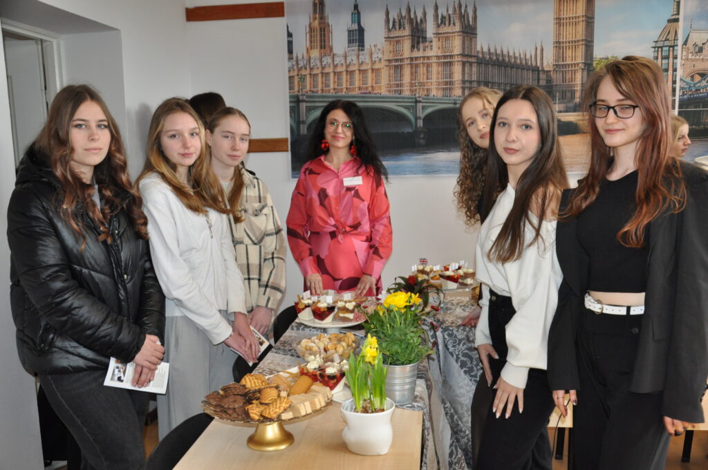 Grupa młodych kobiet stoi przy stole z przekąskami i kwiatami, na tle dużego plakatu z widokiem budynków w Londynie.