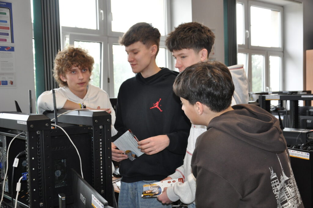 Czterech nastolatków rozmawia i trzyma ulotki przy stanowiskach komputerowych w jasnej sali.