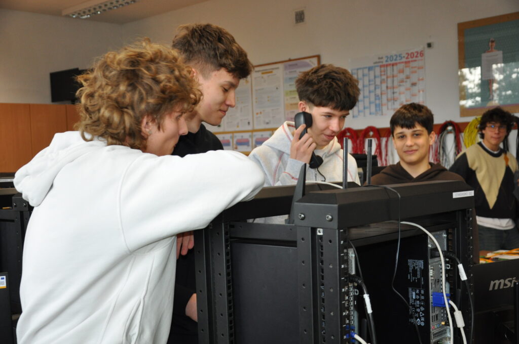 Grupa nastolatków z zaciekawieniem obserwuje sprzęt komputerowy podczas zajęć w pracowni informatycznej.