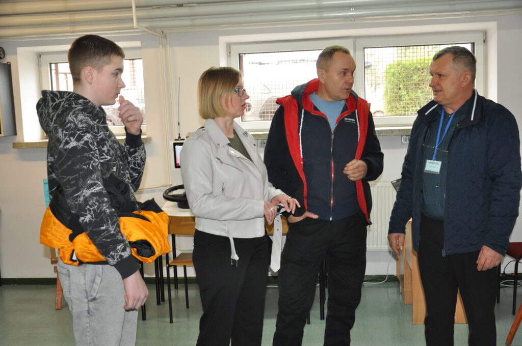 Grupa czterech osób rozmawia w jasnym pomieszczeniu, w tle widać okna i meble.