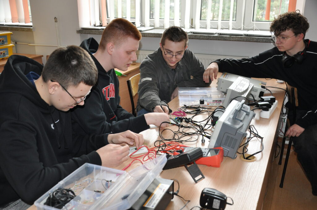 Czterech młodych mężczyzn pracuje wspólnie przy stole, testując i podłączając liczne przewody do urządzeń elektronicznych i przyrządów pomiarowych.