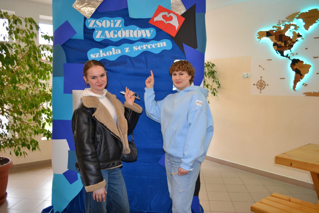 Dwie młode kobiety stoją przed niebieskim banerem z napisem „ZSOiZ Zagórów szkoła z sercem” i wskazują na niego.