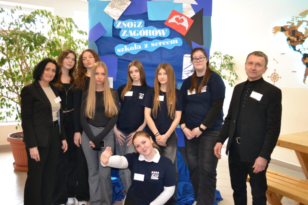 Grupa młodzieży i dwoje dorosłych na tle plakatu z napisem „ZSOiZ Zagórów szkoła z sercem”.