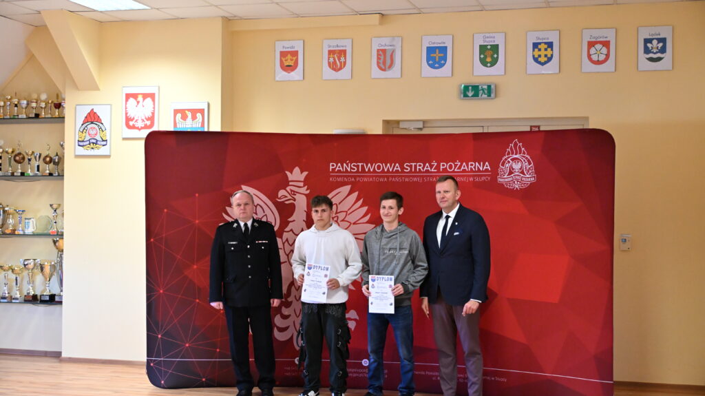 Dwaj młodzi mężczyźni trzymają dyplomy, stojąc między dwoma dorosłymi mężczyznami na tle baneru Państwowej Straży Pożarnej.