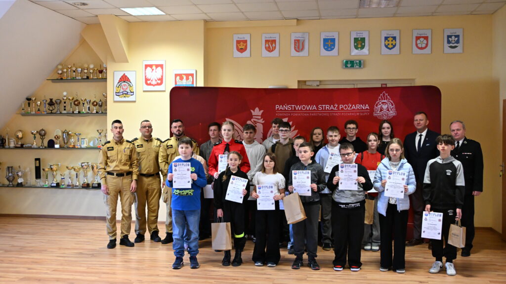 Grupa dzieci i strażaków stoi w pomieszczeniu Komendy Powiatowej Państwowej Straży Pożarnej w Słupcy, a dzieci trzymają dyplomy i torby z upominkami.