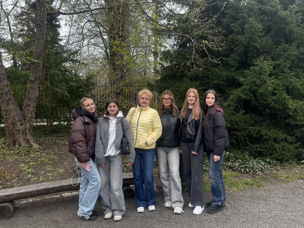 Grupa sześciu kobiet w kurtkach, stojących na zewnątrz na tle drzew i zieleni.