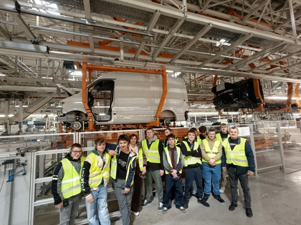 Grupa osób w odblaskowych kamizelkach stoi przed linią montażową, na której zawieszony jest biały samochód dostawczy.