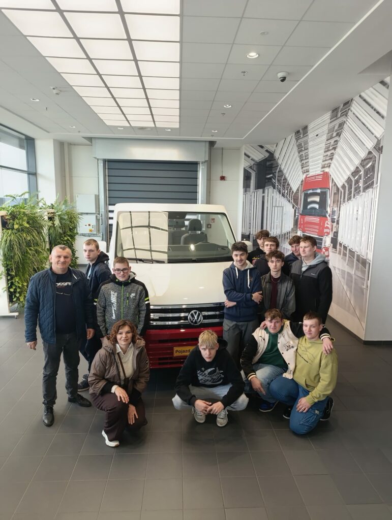 Grupa osób pozująca przed białym samochodem Volkswagen w nowoczesnym wnętrzu z dużymi oknami i roślinami.