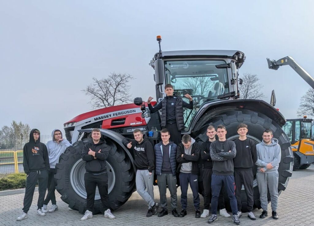 Grupa młodych mężczyzn stoi i pozuje przy dużym czerwono-czarnym ciągniku rolniczym marki Massey Ferguson na utwardzonym placu.