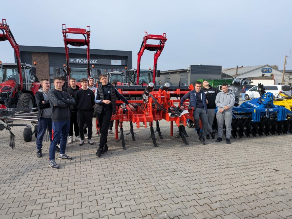 Grupa młodych mężczyzn stoi przy maszynach rolniczych i traktorach na placu przed budynkiem oznaczonym jako EUROMASZ.
