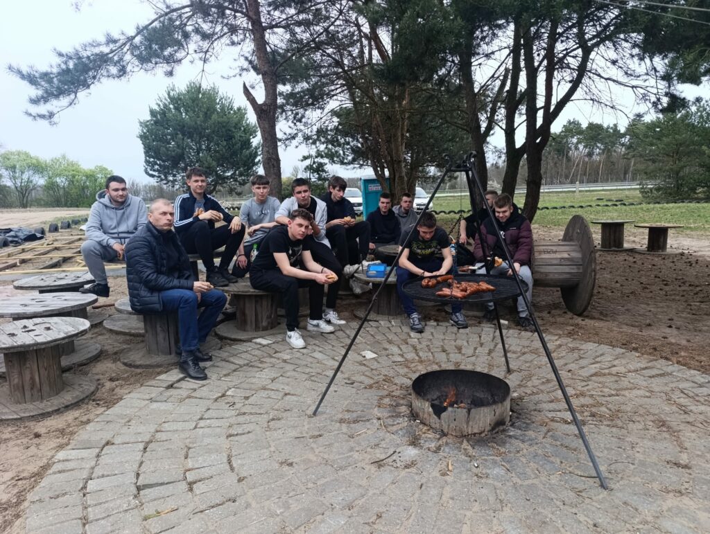 Grupa młodych mężczyzn siedzi na ławkach przy ognisku, nad którym grillowane są kiełbaski.