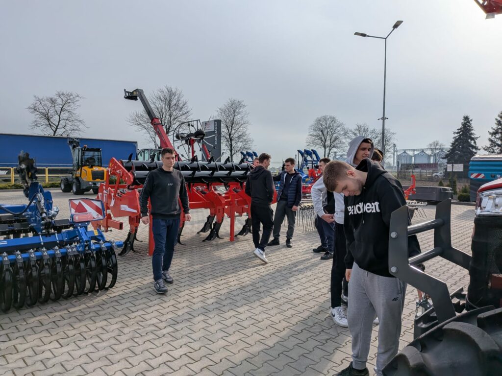 Grupa młodych mężczyzn ogląda i bada różne maszyny rolnicze na zewnątrz.