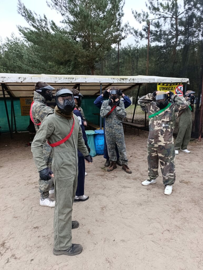 Grupa osób ubranych w kamizelki ochronne i maski do paintballa stoi na polu, przygotowując się do gry.