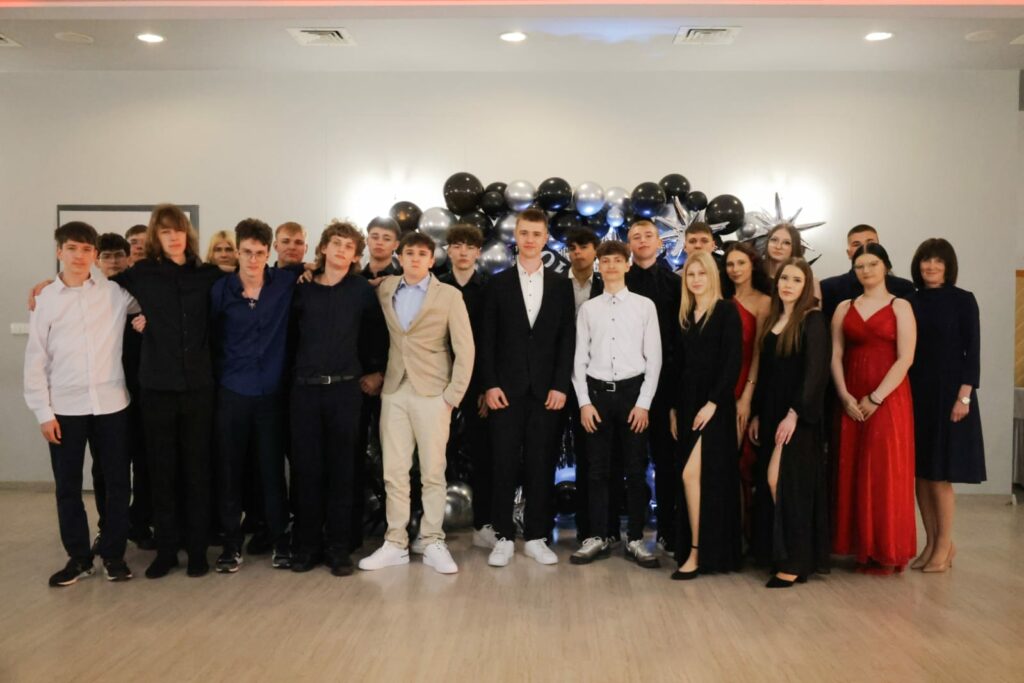 Grupa młodych ludzi ubranych elegancko, ustawionych na tle czarno-srebrnej dekoracji z balonów, pozujących do zbiorowego zdjęcia.