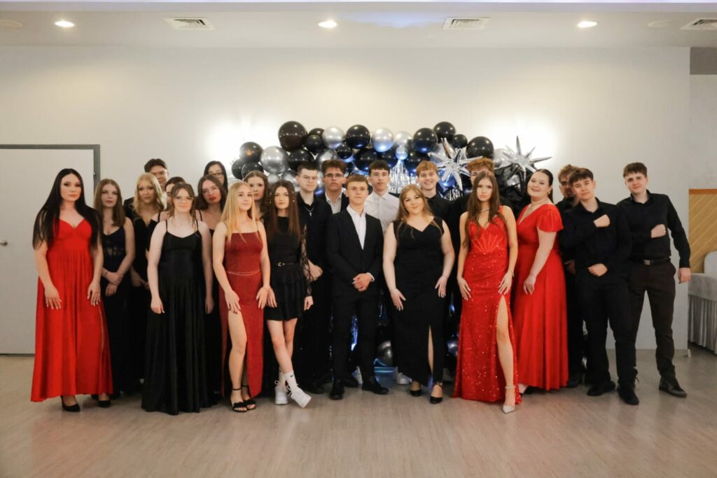 Grupa młodych osób na eleganckim balu w sukienkach i garniturach, na tle dekoracji z czarnych i srebrnych balonów.