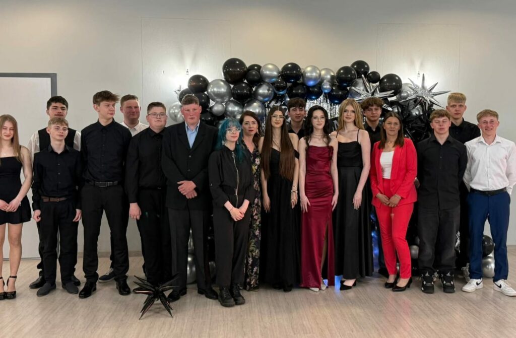 Grupa młodych ludzi oraz dwóch dorosłych, ubranych elegancko, stoi na tle dekoracji z czarno-srebrnych balonów.
