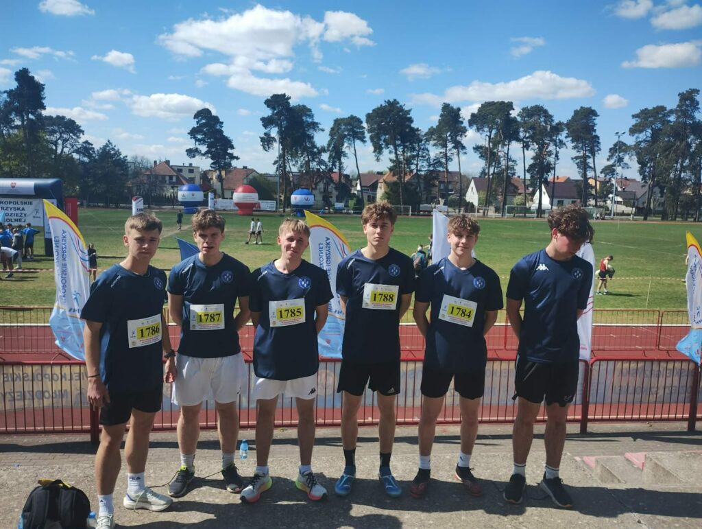 Sześciu młodych mężczyzn w sportowych strojach stoi na tle bieżni lekkoatletycznej podczas zawodów na stadionie.