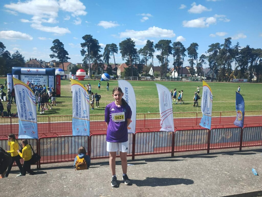 Młoda zawodniczka w fioletowej koszulce i białych spodenkach stoi na trybunach stadionu lekkoatletycznego podczas zawodów sportowych.