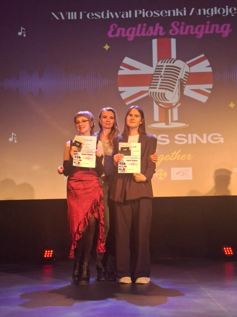 Trzy kobiety na scenie podczas XVIII Festiwalu Piosenki Anglojęzycznej English Singing, dwie z nich trzymają dyplomy i nagrody.