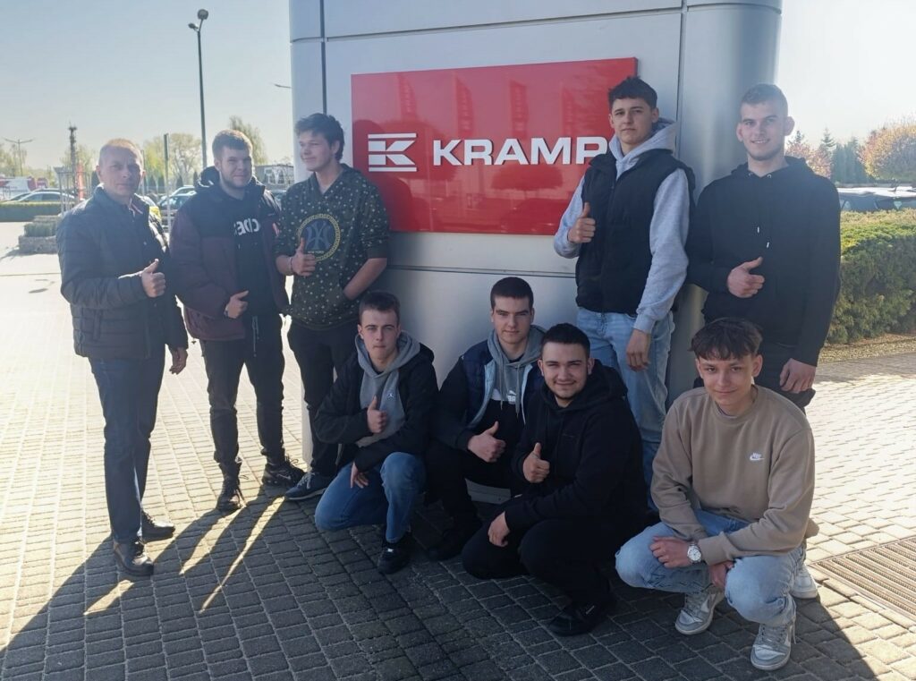 Grupa młodych mężczyzn pozująca przed dużym znakiem firmowym KRAMP, większość pokazuje kciuk w górę.