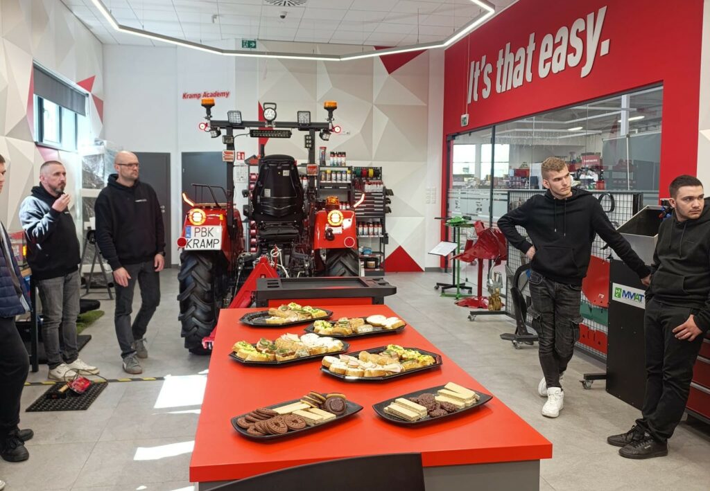 Wewnątrz warsztatu lub szkoleniowego miejsca stoi czerwony traktor z tablicą rejestracyjną, a przy stole z przekąskami stoi kilka osób w czarnych ubraniach, na ścianie widoczny napis "It’s that easy."