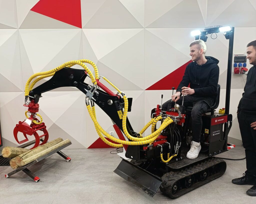 Młody mężczyzna steruje małą koparką hydrauliczną unoszącą drewniane kłody, obok stoi inna osoba obserwująca.