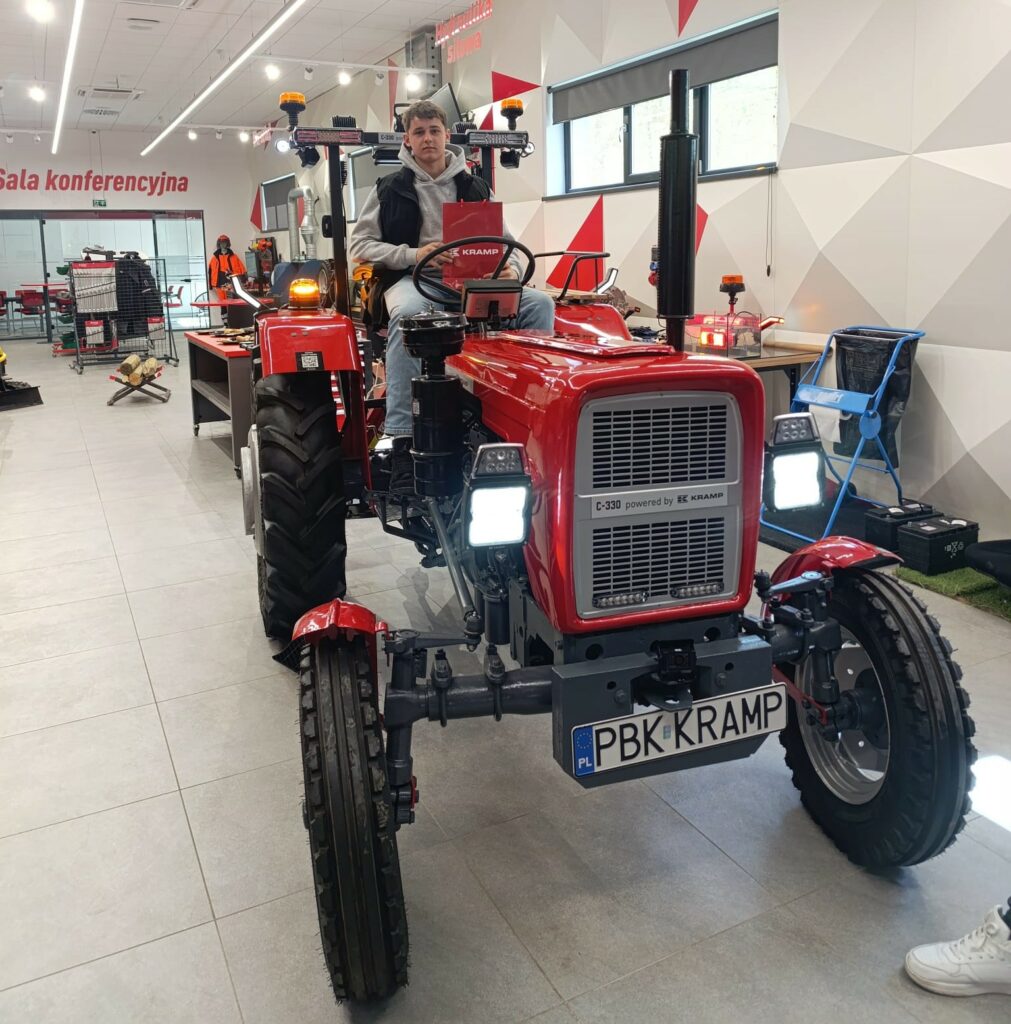 Młody mężczyzna siedzi na czerwonym ciągniku w przestronnym, nowoczesnym wnętrzu z napisem „Sala konferencyjna” na ścianie.