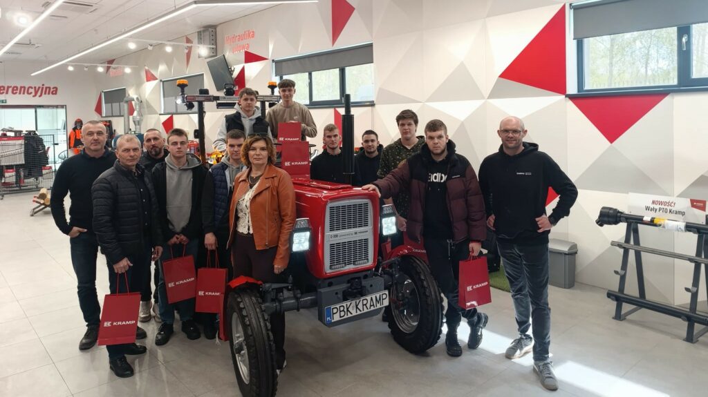 Grupa ludzi stoi wokół czerwonego traktora marki Kramp w nowoczesnym showroomie z motywami czerwono-szarymi na ścianach.