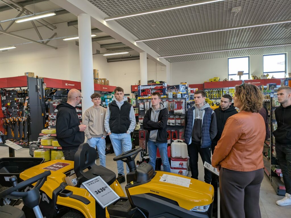 Grupa młodych ludzi w sklepie narzędziowym słucha mężczyzny w czarnej bluzie, stojącego obok żółtych traktorków ogrodowych.