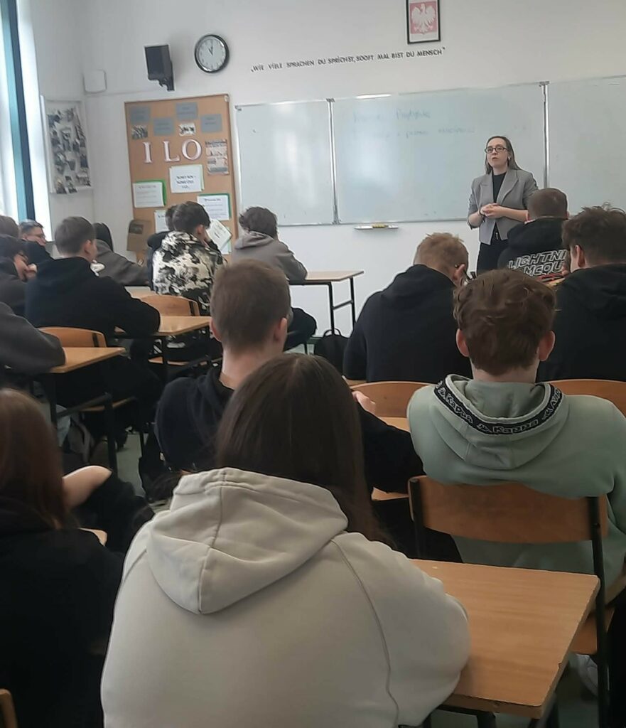 Uczniowie siedzą przy ławkach i słuchają nauczycielki prowadzącej lekcję przy tablicy w klasie szkolnej.