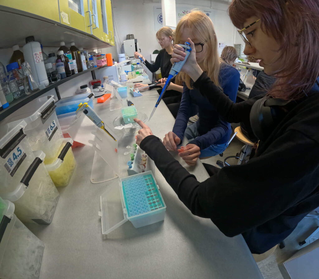 Cztery osoby wykonujące eksperymenty laboratoryjne z użyciem pipet i próbówek przy stole pełnym sprzętu i odczynników.