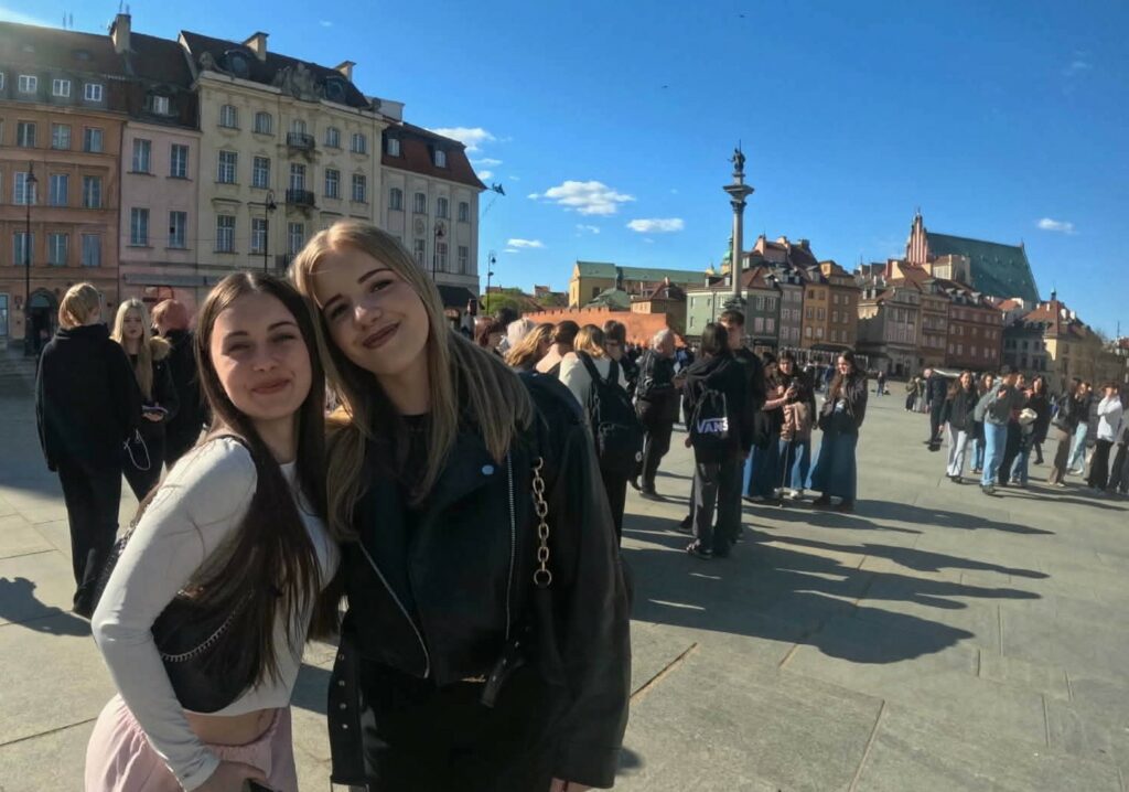 Dwie młode kobiety uśmiechają się na tle tłumu ludzi stojących na placu z zabytkowymi kamienicami i kolumną z figurą w oddali.